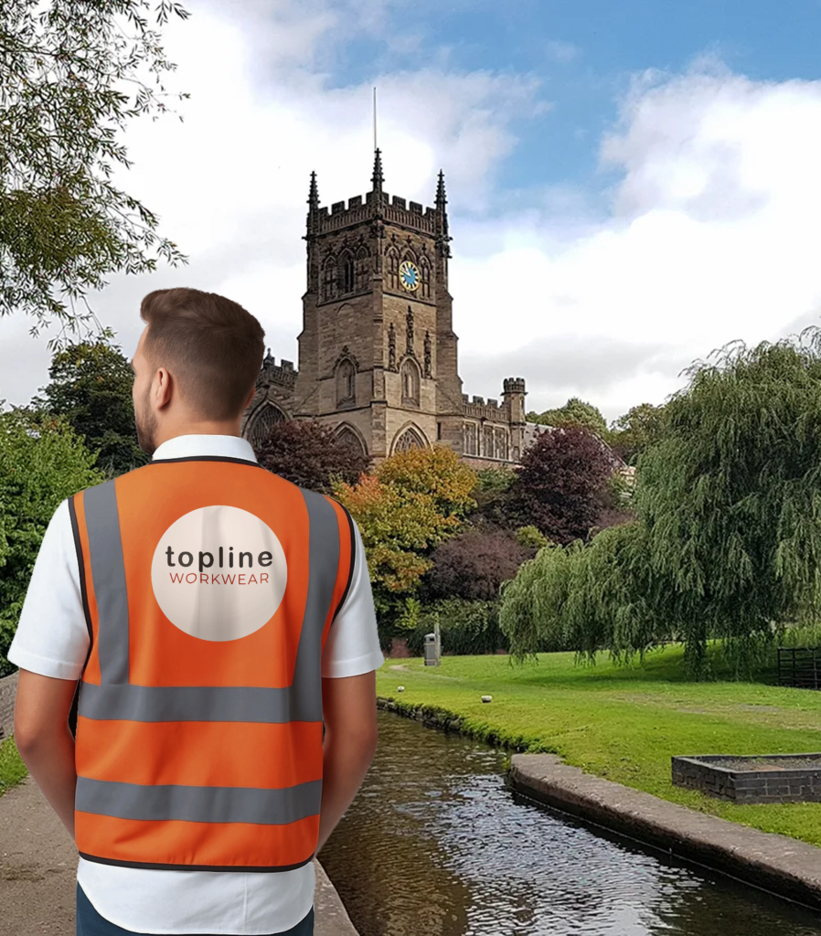 Person seen from behind in a bright orange high‑visibility vest with'topline' logo, standing in a park with a historic clock tower