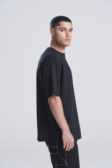 Man in a loose black T-shirt and dark jeans, standing in profile against a light studio backdrop.