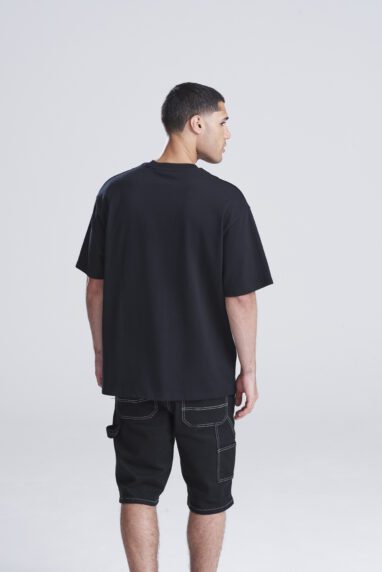 Man seen from the back wearing a loose black t-shirt and knee-length black shorts in a studio setting