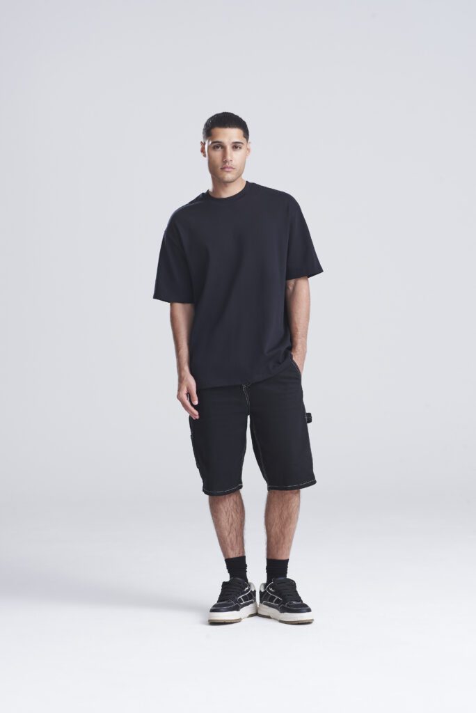 Man model wearing a loose black T-shirt, black knee-length shorts, black socks and sneakers in a studio backdrop.