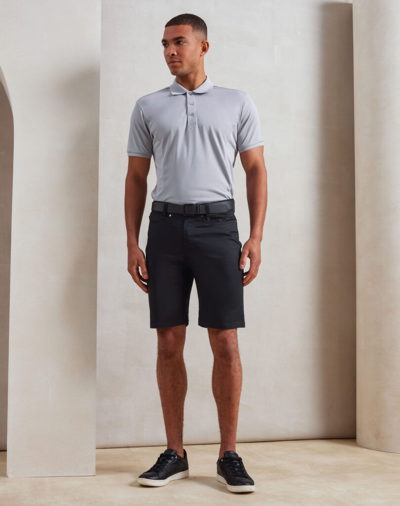 Man standing in a neutral studio setting wearing a light gray polo shirt, black shorts, and black sneakers with white soles.