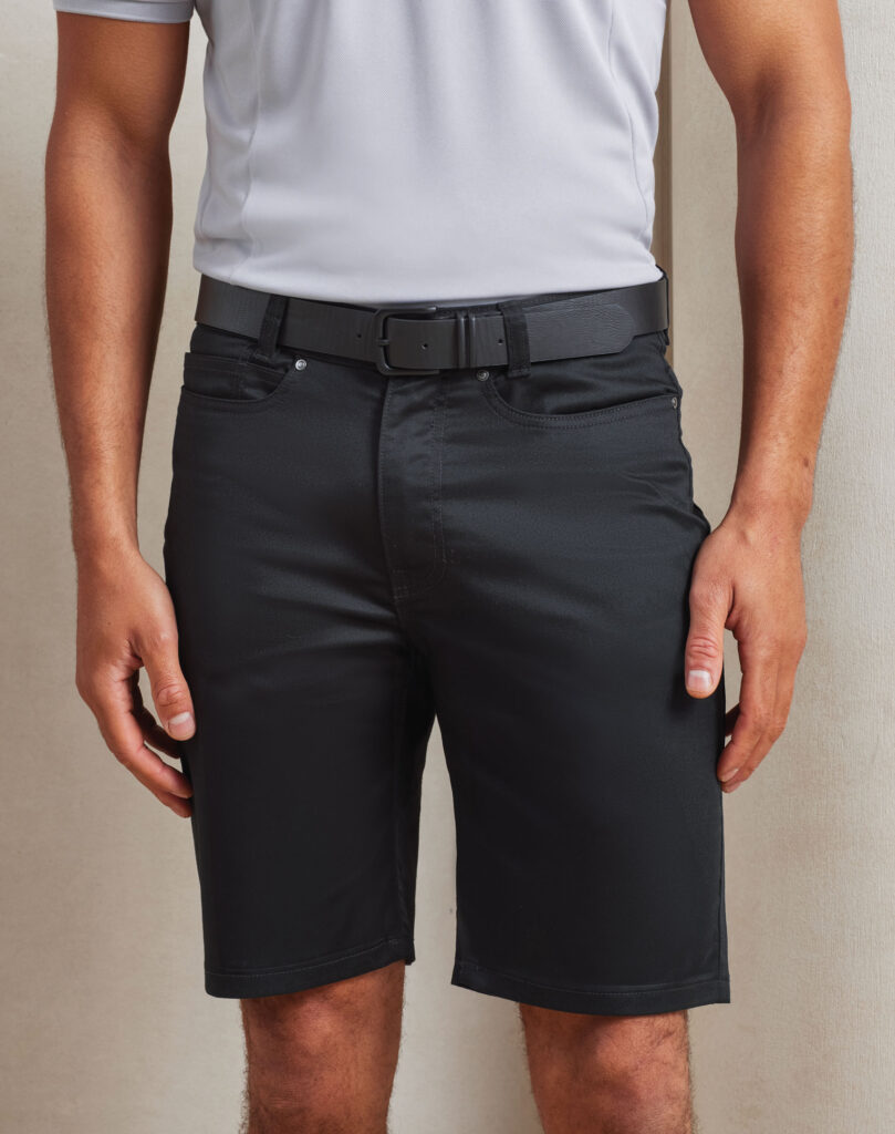 Man wearing a white shirt with black shorts and a black belt, standing against a beige wall (midsection to knees).