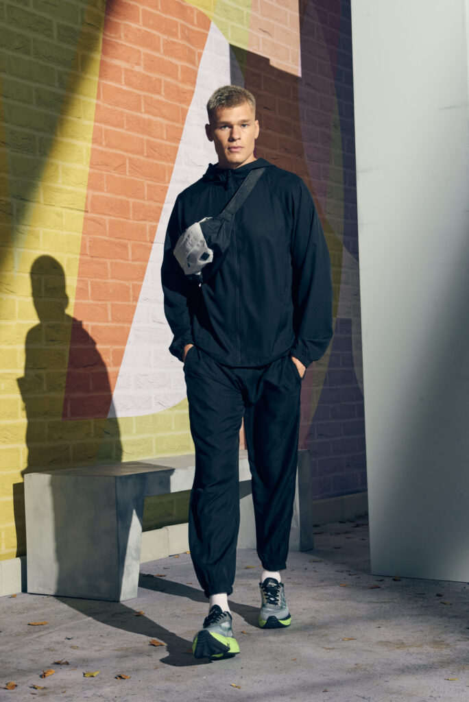 JC281_JC282_Jet_Black_18mths_start_31_Dec_2024_1096 - Topline Workwear Man in a black tracksuit stands against a colorful mural, with a crossbody bag over his chest.