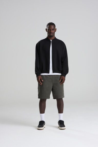 JH124_DEEP_BLACK_DEC2025-DEC2027_109-382x572 - Topline Workwear Adult man with dark skin standing in a studio wearing a black zip-up jacket, white T-shirt, olive shorts, white socks, and black sneakers.